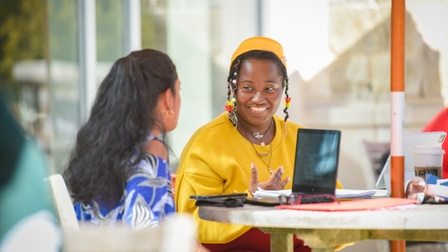 Two female students sitting at table on laptops talking